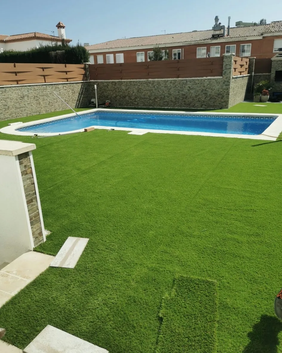 Franja de césped artificial junto a una piscina, con un seto alto recortado detrás