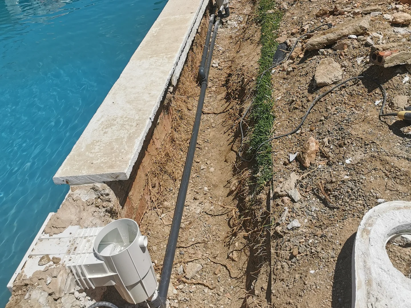 Esquina de una piscina con el skimmer desmontado en el suelo y la zanja abierta a lo largo del bordillo con la tubería a la vista